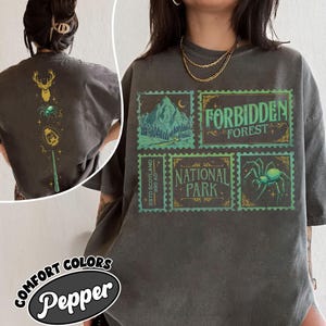 May include: A charcoal gray t-shirt featuring a front graphic of four postage stamp-style boxes with green and gold illustrations of a forest, a spider, and text reading Forbidden Forest and National Park. The back shows a vertical line of icons.