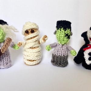 Puede incluir: Cuatro marionetas de dedo de punto vestidas como personajes de Halloween: una bruja, una momia, el monstruo de Frankenstein y un vampiro. La bruja lleva un sombrero negro y un vestido morado, la momia es blanca, el monstruo de Frankenstein es verde y morado, y el vampiro es negro con una capa roja.