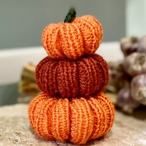 May include: A stack of three crocheted pumpkins in shades of orange. The pumpkins are stacked on top of each other, with the smallest pumpkin on top. The pumpkins are made with a textured stitch, giving them a rustic look.