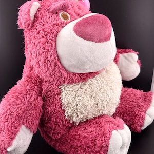 May include: Pink plush toy bear with a white belly and a green leaf on its head. The bear is sitting with its legs crossed.