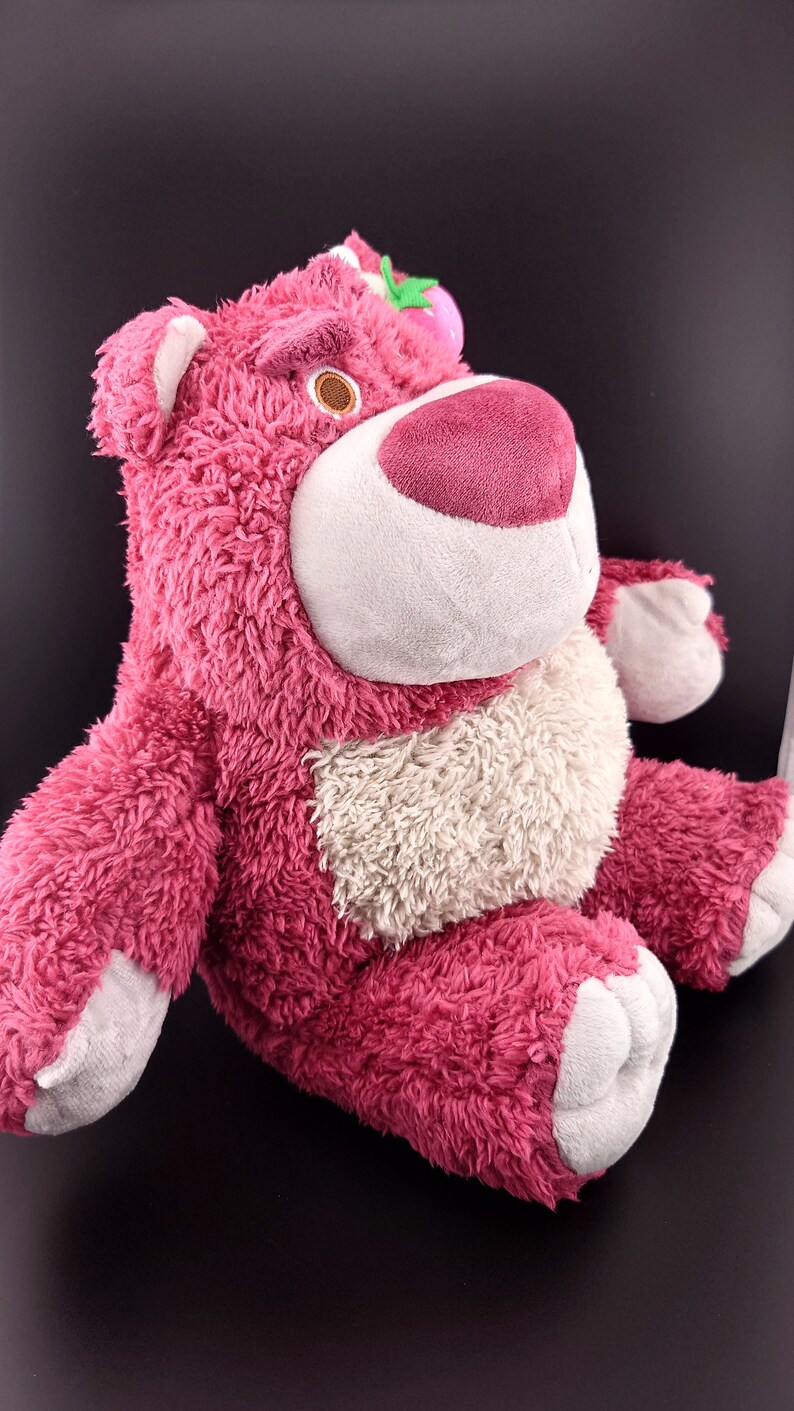 May include: Pink plush toy bear with a white belly and a green leaf on its head. The bear is sitting with its legs crossed.