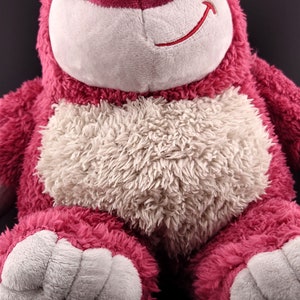 May include: A plush pink and white teddy bear with a strawberry on its head. The bear has a white nose and a stitched smile.
