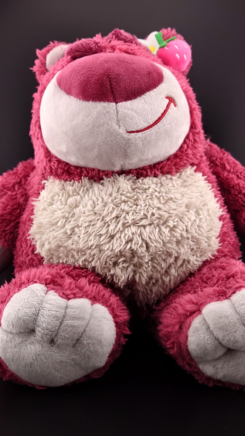 May include: A plush pink and white teddy bear with a strawberry on its head. The bear has a white nose and a stitched smile.