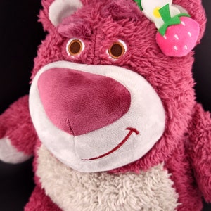 May include: Pink plush toy bear with a white face and a smiling mouth. The bear has a white fur collar and a strawberry flower on its head.