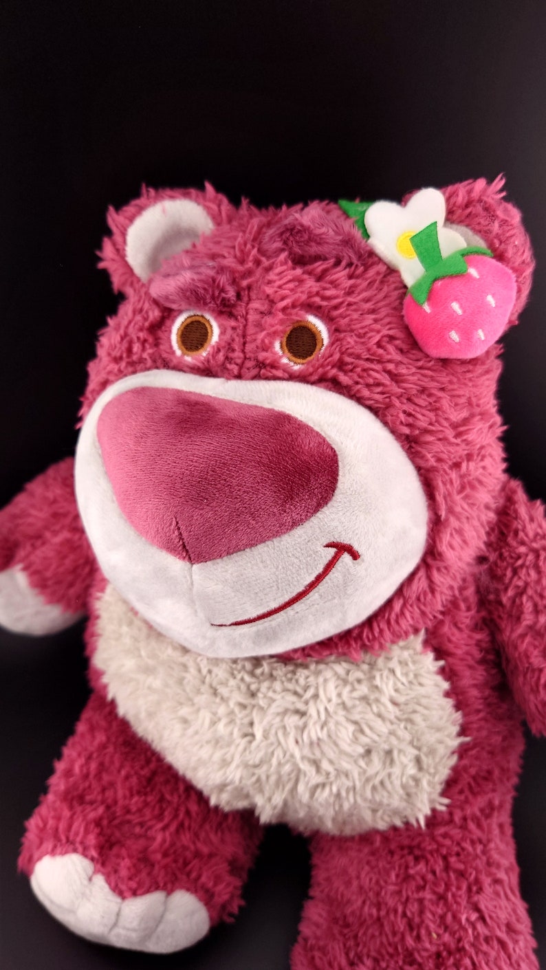 May include: Pink plush toy bear with a white face and a smiling mouth. The bear has a white fur collar and a strawberry flower on its head.