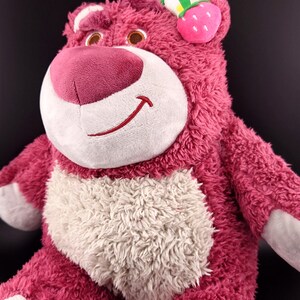 May include: Pink plush toy bear with a white belly and a strawberry on its head. The bear has a friendly smile and is made of soft, fuzzy material.