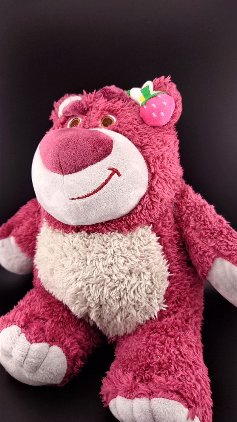 May include: Pink plush toy bear with a white belly and a strawberry on its head. The bear has a friendly smile and is made of soft, fuzzy material.
