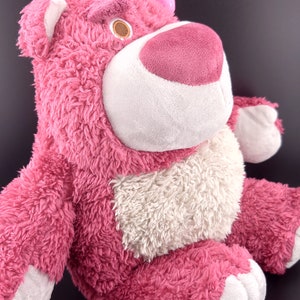 May include: Pink plush toy bear with a white belly and a pink nose. The bear has a green leaf and a pink flower on its head.
