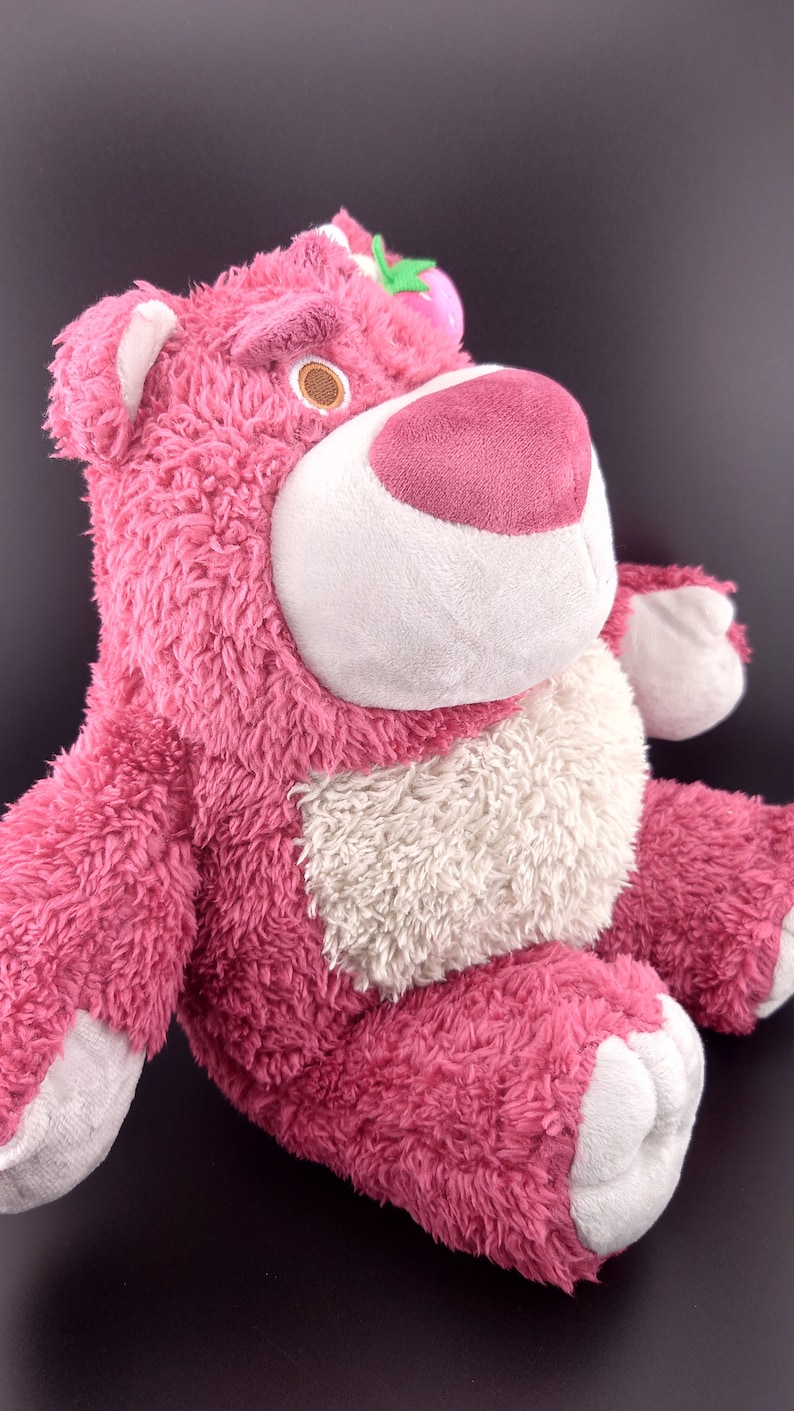 May include: Pink plush toy bear with a white belly and a pink nose. The bear has a green leaf and a pink flower on its head.