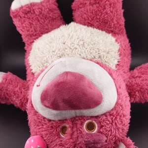 May include: Pink plush toy bear with a white belly and a strawberry and flower on its head.