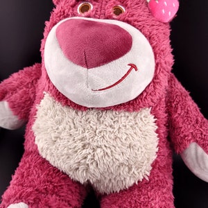 May include: Pink plush toy bear with a white face and a white belly. The bear has a flower and a strawberry on its head.