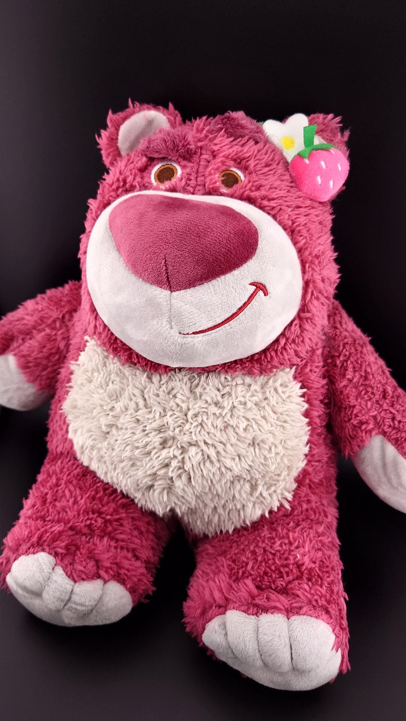 May include: Pink plush toy bear with a white face and a white belly. The bear has a flower and a strawberry on its head.