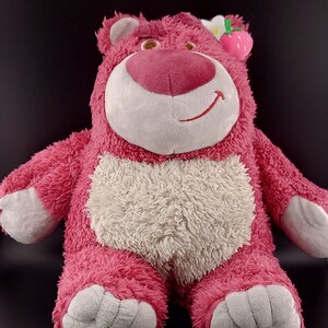 May include: Pink plush toy bear with a white belly and a strawberry on its head. The bear has a friendly expression and is sitting with its legs crossed.