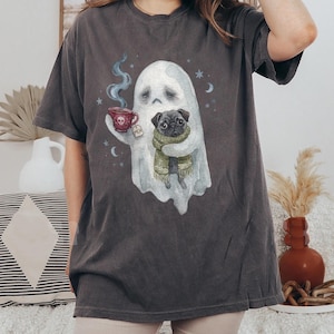 May include: A charcoal grey t-shirt featuring a graphic illustration of a white ghost holding a red skull-patterned teacup with a tag labelled "MEH" and embracing a black pug wearing a green knit scarf, surrounded by small blue stars and moons.