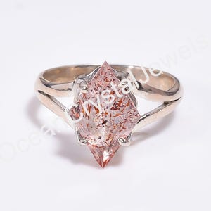 May include: A silver ring featuring a marquise-cut, light pink gemstone with internal inclusions. The gemstone is set in a split-shank band. The ring is displayed against a white background, highlighting the gemstone's color and clarity.