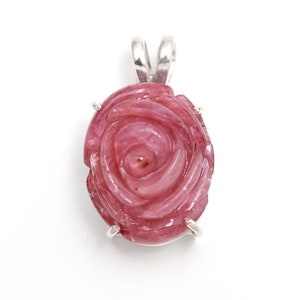May include: A pink gemstone pendant carved in the shape of a rose. The pendant has a silver bail and four silver prongs. The rose is intricately detailed, and the gemstone has a polished finish. The background is white.