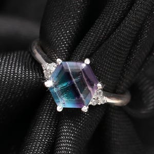 May include: A silver ring with a hexagonal gemstone in shades of blue, green, and purple. The gemstone is flanked by small, clear stones. The ring is set against a dark, textured backdrop.