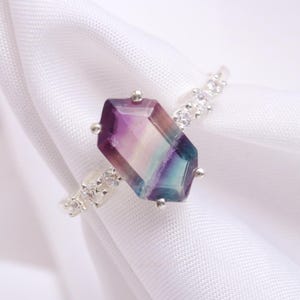 Peut inclure: Une bague en argent avec une grande pierre de fluorite facetée multicolore. La bague est sertie de petites pierres de zircone cubique transparentes.