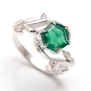 Puede incluir: Un anillo de plata con una piedra preciosa verde engastada en forma hexagonal. El anillo presenta un delicado diseño de hojas y enredaderas.