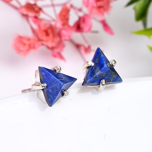 May include: A pair of triangular lapis lazuli stud earrings. Each earring has a deep blue, faceted gemstone set in a silver metal setting. The earrings are displayed on a white surface, with pink flowers in the background.