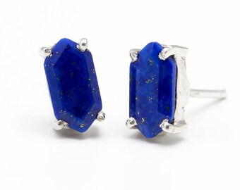 May include: A pair of stud earrings featuring rectangular, faceted, deep blue lapis lazuli stones. The stones are set in a silver-colored metal with four small prongs holding each stone. The earrings are displayed against a white background.