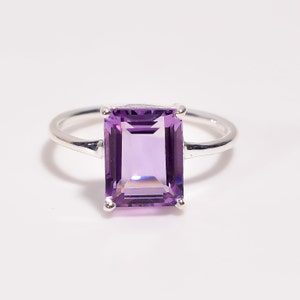 May include: A silver ring featuring a large, rectangular, faceted amethyst gemstone. The amethyst is a vibrant purple color, and the ring has a simple, elegant design. The gemstone is approximately 0.75 inches long.