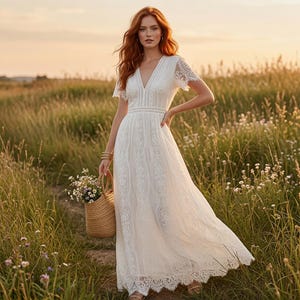 May include: A floor-length white lace dress featuring short sleeves, a V-neckline, and a fitted waist with intricate vertical embroidery patterns. The dress is paired with a woven straw basket containing a small bouquet of wildflowers.