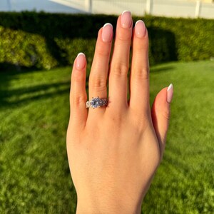 May include: A close-up of a hand wearing a silver ring with a large, clear, round gemstone surrounded by smaller gemstones. The hand has a French manicure with white tips.