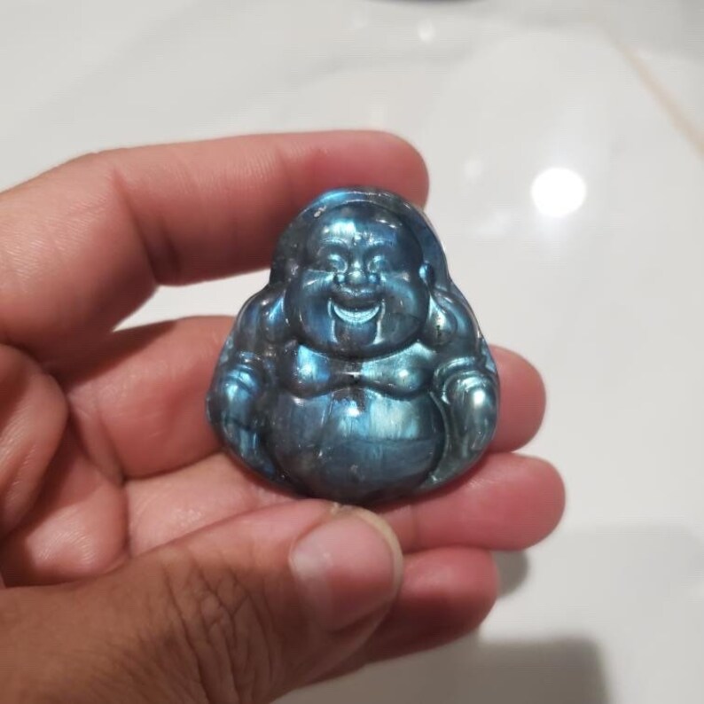 May include: A small, smiling Buddha figurine carved from a blue-gray stone. The Buddha has a rounded belly and a serene expression. The stone has iridescent flashes of blue and green.