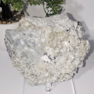 May include: A raw, natural aquamarine crystal cluster. The stone features a pale blue hue with a crystalline structure, set against a matrix of white and gray minerals. The rough texture and natural form are prominent.