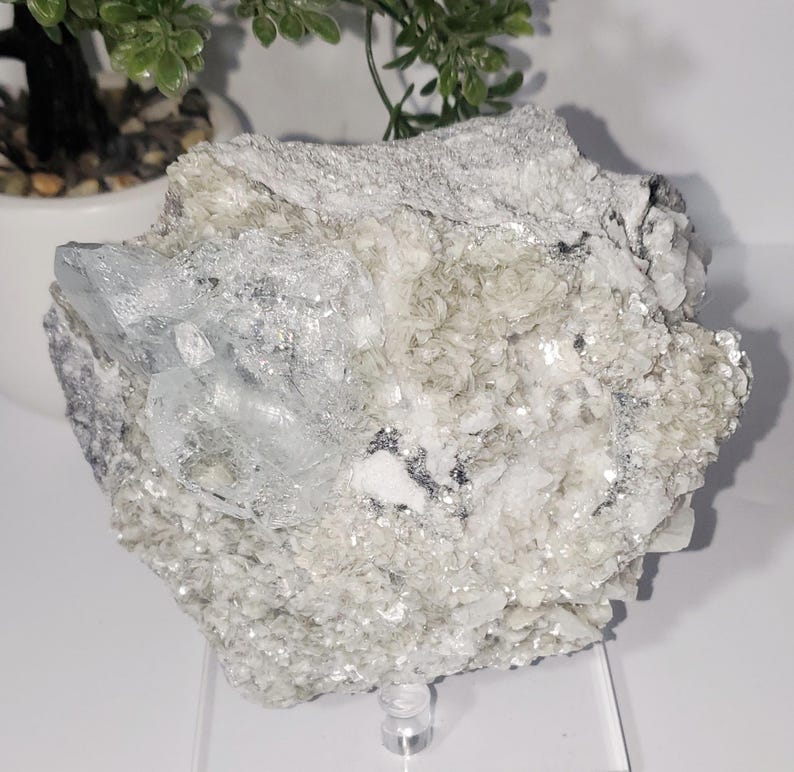 May include: A raw, natural aquamarine crystal cluster. The stone features a pale blue hue with a crystalline structure, set against a matrix of white and gray minerals. The rough texture and natural form are prominent.