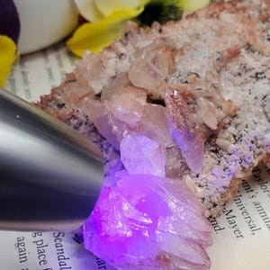 K&ouml;nnte beinhalten: Ein Cluster aus rosa und wei&szlig;en Kristallen, die von einem UV-Licht beleuchtet werden und einen leuchtend violetten Schein erzeugen. Die Kristalle sind auf einem Buch mit sichtbarem Text angeordnet.