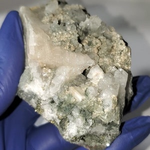 May include: A cluster of white quartz crystals embedded in a matrix of green and gray rock. The crystals are translucent and have a glassy luster.