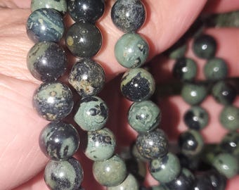 May include: Three beaded bracelets with dark green and black spherical beads. The beads have a mottled appearance, with variations in color and texture. The bracelets are held in a hand, showcasing the jewelry's design and color variations.