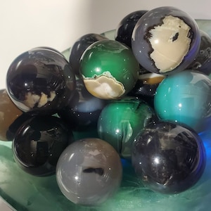 May include: A collection of polished gemstone spheres in various colors, including green, gray, black, and brown. The spheres are displayed in a clear, shallow bowl with a blue-green hue. The gemstones have unique patterns and textures.
