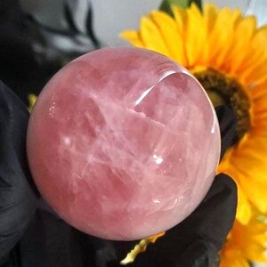 May include: A large, pink rose quartz sphere with a smooth, polished surface. The sphere is held in a hand, with a yellow sunflower in the background.