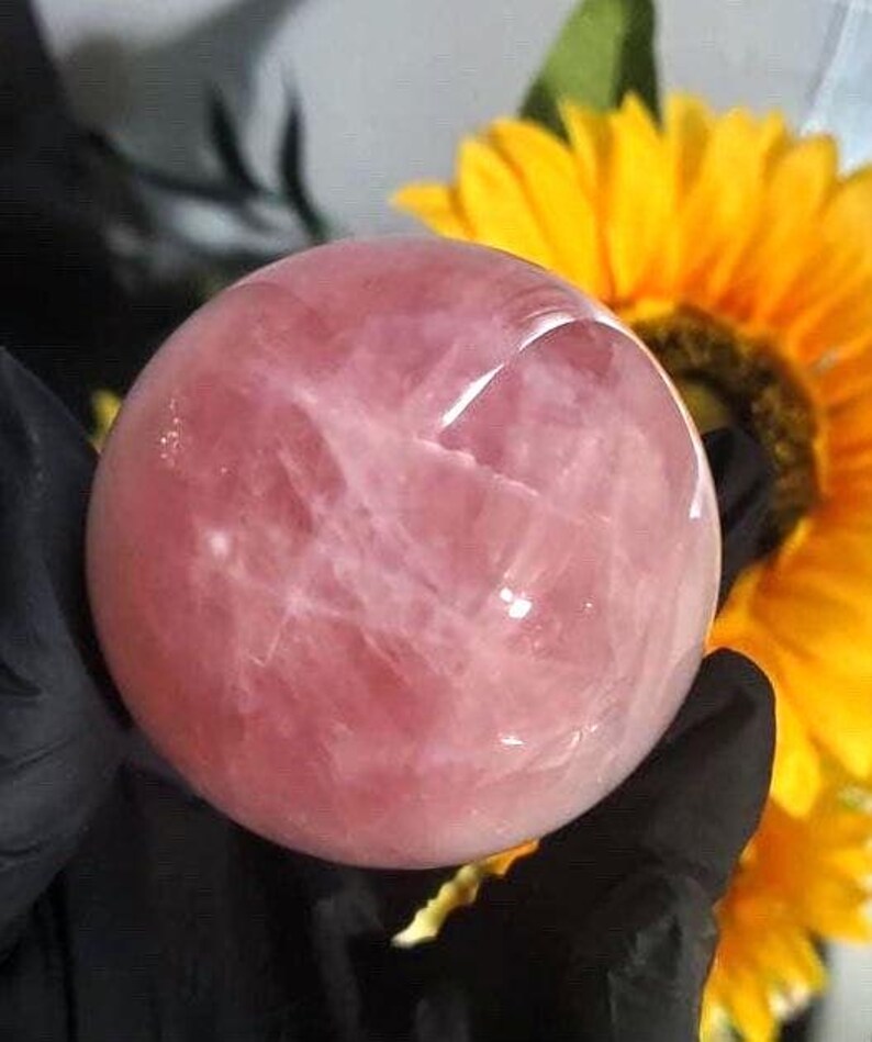 May include: A large, pink rose quartz sphere with a smooth, polished surface. The sphere is held in a hand, with a yellow sunflower in the background.