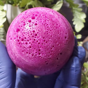 May include: A close-up of a magenta sphere with a porous texture. The sphere is held by a gloved hand. The surface of the sphere is covered in various sized holes. Green foliage is visible in the background.