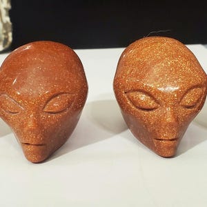 May include: Four alien head sculptures carved from different types of stone. The sculptures are all about 3 inches tall and have a similar design with large eyes and a small mouth. The sculptures are in various colors, including purple, brown, orange, and green.