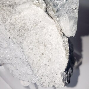 May include: A close-up of a raw, crystalline mineral specimen. The stone features a mix of white, gray, and clear crystal formations. The rough texture and natural shapes are visible, with light reflecting off the surfaces.