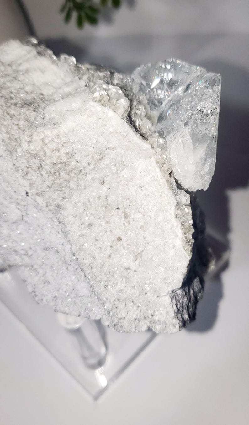 May include: A close-up of a raw, crystalline mineral specimen. The stone features a mix of white, gray, and clear crystal formations. The rough texture and natural shapes are visible, with light reflecting off the surfaces.