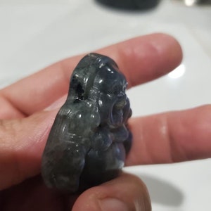 May include: A small, carved figurine of a laughing Buddha. The sculpture is a dark, iridescent gray-blue color with visible details of the Buddha's face and body. The figurine is held in a person's hand.