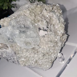 May include: A cluster of raw, uncut crystals in shades of white, gray, and clear. The crystals have a rough, textured surface with a sparkling, reflective quality. The overall appearance is natural and organic, with a focus on the mineral's unique form.