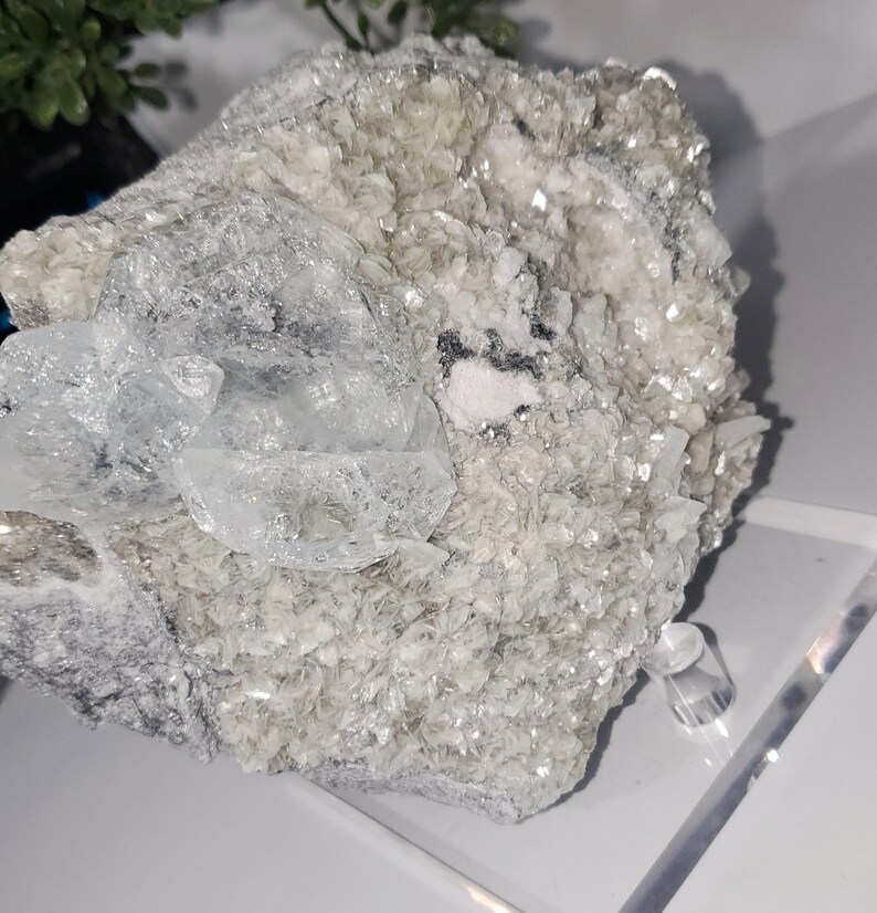 May include: A cluster of raw, uncut crystals in shades of white, gray, and clear. The crystals have a rough, textured surface with a sparkling, reflective quality. The overall appearance is natural and organic, with a focus on the mineral's unique form.