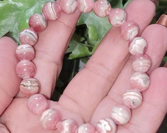 May include: A stretch bracelet composed of round, polished rhodochrosite beads. Each bead displays a unique pattern of swirling pink, white, and grey bands, creating a natural, banded appearance throughout the circular arrangement of the gemstone jewelry.