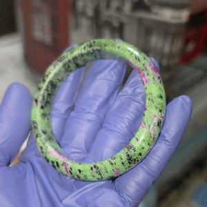 May include: A polished gemstone bangle bracelet. The bracelet is a vibrant green color with black and pink inclusions. The smooth, circular design is held in a gloved hand, showcasing the natural stone's unique patterns and colors.