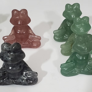 May include: Four small frog figurines carved from green, pink, and gray stone. The frogs are in a seated meditation pose with their eyes closed.