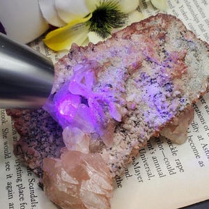 K&ouml;nnte beinhalten: Ein Cluster aus rosa und wei&szlig;en Kristallen auf einem Stein. Die Kristalle werden mit UV-Licht beleuchtet, wodurch sie in einem leuchtenden Lila leuchten.