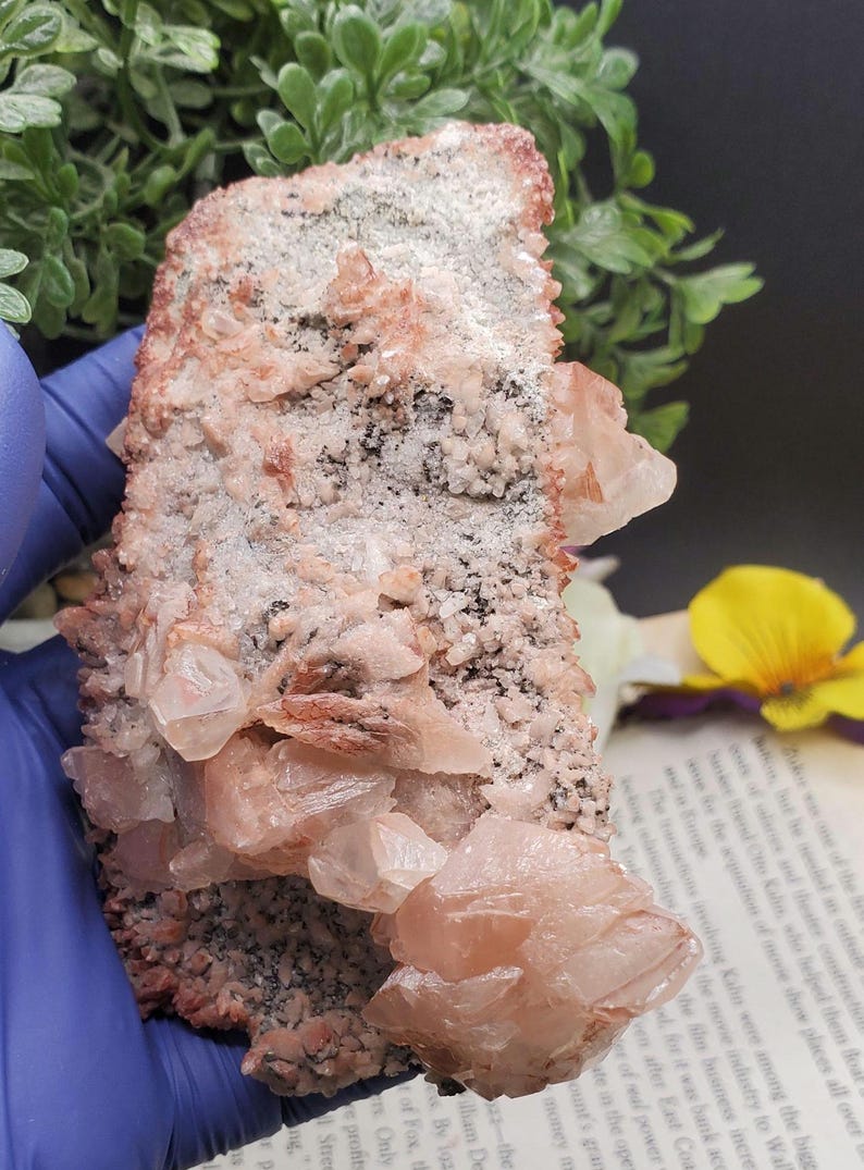 K&ouml;nnte beinhalten: Ein Cluster aus rosa und wei&szlig;en Calcitkristallen mit einer rauen, grauen und braunen Matrix. Die Kristalle sind durchscheinend und haben einen leicht schillernden Glanz.