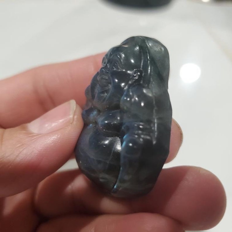 May include: A carved, blue-gray gemstone figurine of a seated Buddha. The figure has a rounded belly and a serene expression. The stone has a polished, reflective surface, with subtle color variations. The Buddha is held in a hand.